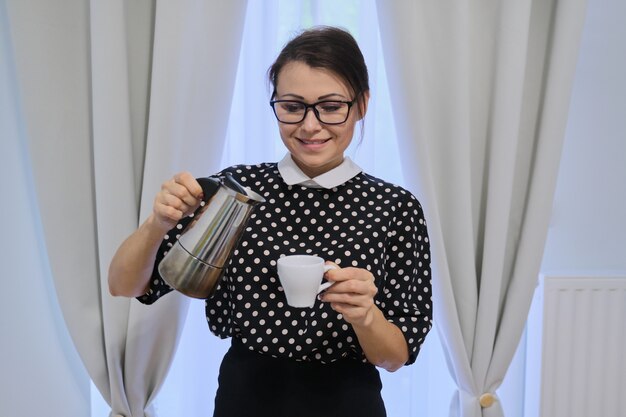 Dona de casa adulta fazendo café, mulher segurando a cafeteira e a xícara, em pé perto da janela com cortinas, hora do intervalo para beber café