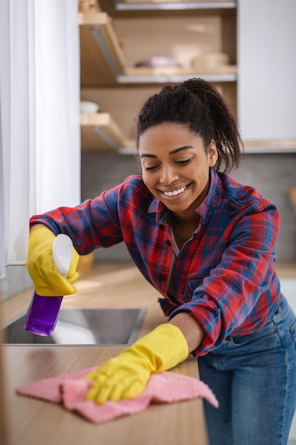 Foto dona de casa afro-americana milenar sorridente em luvas de borracha com spray e pano limpa sujeira na mesa