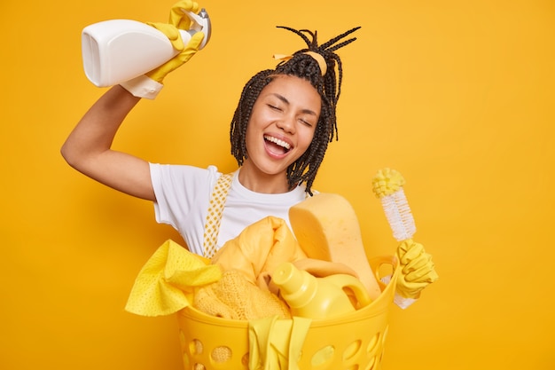 Dona de casa alegre se diverte detém detergente e escova danças contra fundo amarelo cesta de roupa gosta de limpar a casa expressa emoções positivas. conceito de tarefas domésticas de limpeza