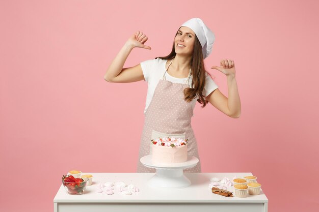 Dona de casa chef feminino cozinhar confeiteiro ou padeiro em avental branco t-shirt, chapéu de chefs de toque cozinhar bolo ou cupcake na mesa isolado no fundo rosa pastel no estúdio. Mock up conceito de comida de espaço de cópia.