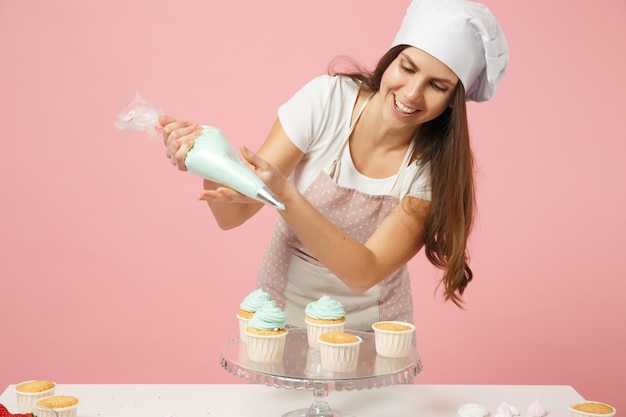 Foto dona de casa chef feminino cozinhar confeiteiro ou padeiro em t-shirt avental branco, chapéu de chefs de toque decorando cupcake de bolo na mesa isolada em fundo rosa pastel no estúdio. mock up conceito de comida de espaço de cópia.