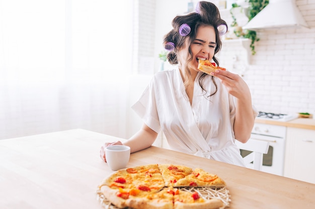 Dona de casa com fome jovem sentar na cozinha e comer pizza. Morda uma fatia de comida. Sozinho no quarto. Governanta aproveitar a vida sem trabalho. Segure o copo nas mãos.