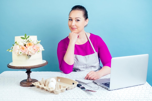 Dona de casa confeiteira (confeiteira) empresária trabalha com um laptop ao lado de bolo de casamento branco de duas camadas com flores frescas em uma mesa com uma toalha de renda em estúdio em um fundo azul