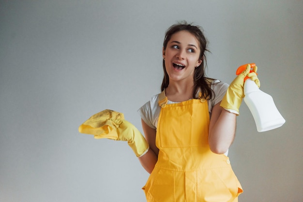 Dona de casa de uniforme amarelo em pé com spray de limpeza para janelas dentro de casa Concepção de renovação de casa