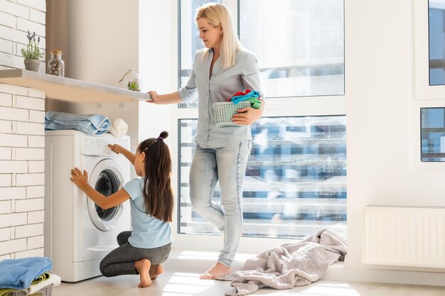 Dona de casa feliz e sua filha com roupa perto da máquina de lavar