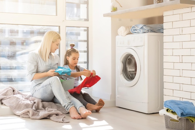 Dona de casa feliz e sua filha com roupa perto da máquina de lavar