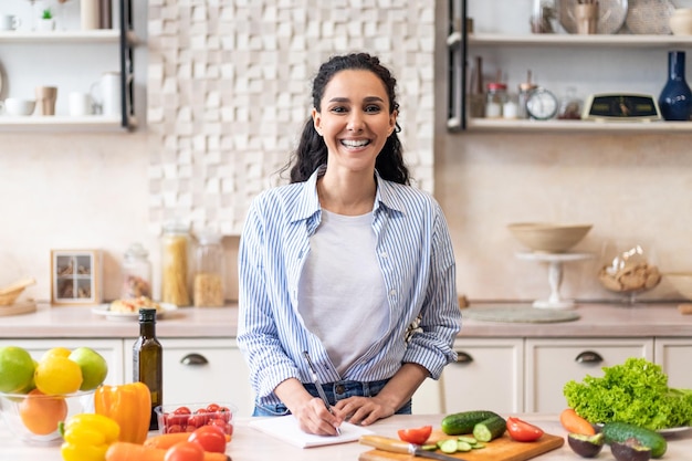 Dona de casa feliz escrevendo novas receitas de comida em seu caderno em pé no interior da cozinha doméstica e