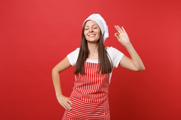 Dona de casa feminina chef cozinheira ou padeiro em avental listrado de camiseta branca, chapéu de chefs toque isolado no fundo da parede vermelha. mulher bonita e calma sorridente, fazendo sinal de prazer bom gosto. mock-se o conceito de espaço de cópia.