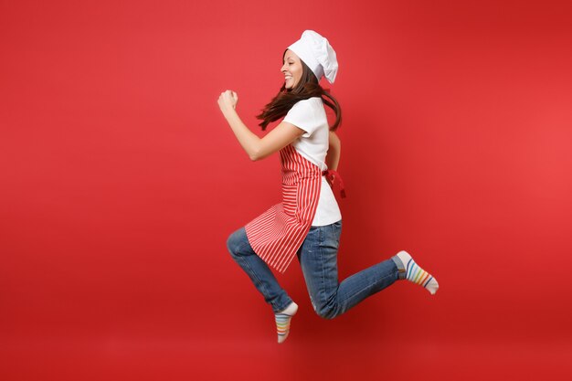 Dona de casa feminina chef cozinheira ou padeiro em avental listrado de camiseta branca, chapéu de chefs toque isolado no fundo da parede vermelha. Mulher governanta de retrato de corpo inteiro pulando alto. Mock-se o conceito de espaço de cópia.