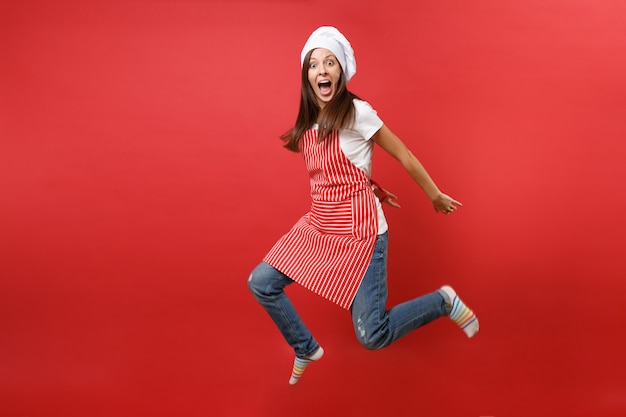 Dona de casa feminina chef cozinheira ou padeiro em avental listrado de camiseta branca, chapéu de chefs toque isolado no fundo da parede vermelha. Mulher governanta de retrato de corpo inteiro pulando alto. Mock-se o conceito de espaço de cópia.