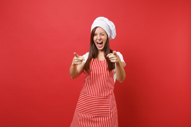 Foto dona de casa feminina chef cozinheira ou padeiro no avental listrado, camiseta branca, chapéu de chefs toque isolado no fundo da parede vermelha. câmera de dedos apontando mulher linda governanta. mock-se o conceito de espaço de cópia.