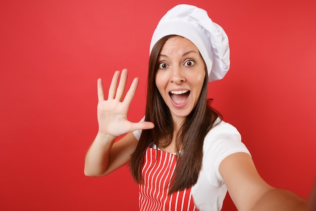 Dona de casa feminina chef cozinheira ou padeiro no avental listrado, camiseta branca, chapéu de chefs toque isolado no fundo da parede vermelha. Feche a mulher dona de casa fazendo tirar uma foto de selfie. Mock-se o conceito de espaço de cópia.