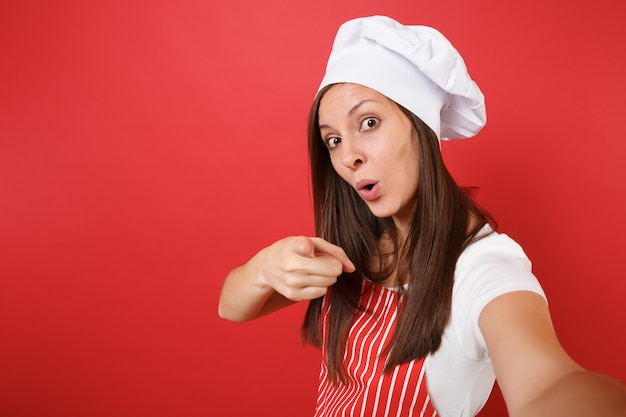 Dona de casa feminina chef cozinheira ou padeiro no avental listrado, camiseta branca, chapéu de chefs toque isolado no fundo da parede vermelha. Feche a mulher dona de casa fazendo tirar uma foto de selfie. Mock-se o conceito de espaço de cópia.