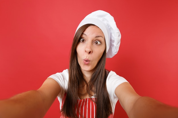 Foto dona de casa feminina chef cozinheira ou padeiro no avental listrado, camiseta branca, chapéu de chefs toque isolado no fundo da parede vermelha. feche a mulher dona de casa fazendo tirar uma foto de selfie. mock-se o conceito de espaço de cópia.