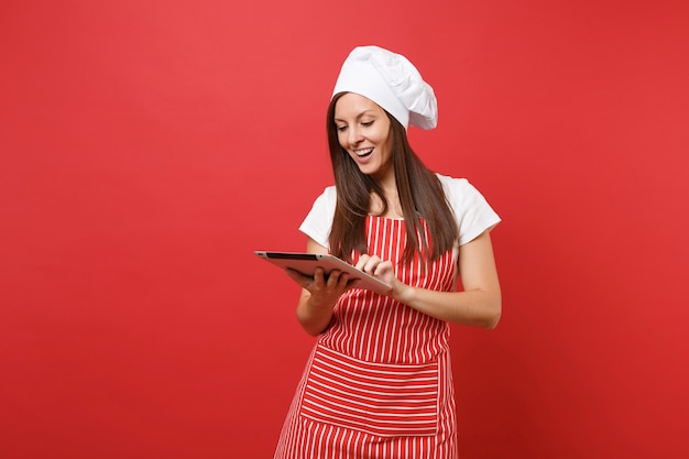 Dona de casa feminina chef cozinheira ou padeiro no avental listrado, camiseta branca, chapéu de chefs toque isolado no fundo da parede vermelha. Mulher divertida governanta procurando receita no tablet pc. Mock-se o conceito de espaço de cópia.