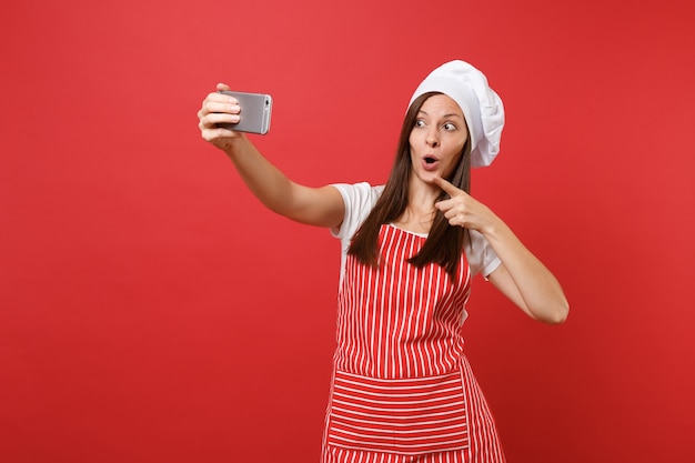 Dona de casa feminina chef cozinheira ou padeiro no avental listrado, camiseta branca, chapéu de chefs toque isolado no fundo da parede vermelha. Mulher divertida sorridente fazendo selfie filmado no celular. Mock-se o conceito de espaço de cópia.