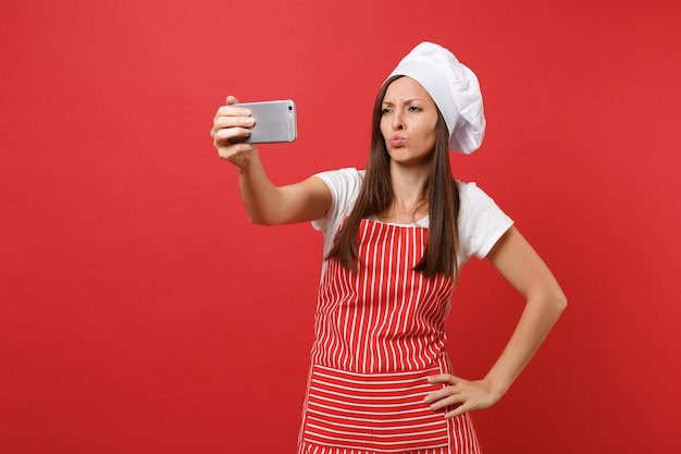 Dona de casa feminina chef cozinheira ou padeiro no avental listrado, camiseta branca, chapéu de chefs toque isolado no fundo da parede vermelha. Mulher divertida sorridente fazendo selfie filmado no celular. Mock-se o conceito de espaço de cópia.