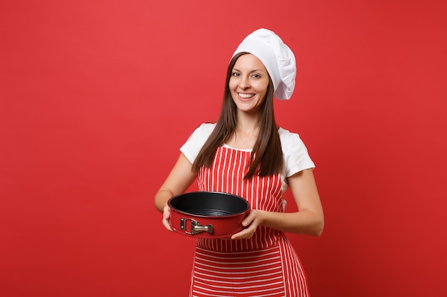 Dona de casa feminina chef cozinheira ou padeiro no avental listrado, camiseta branca, chapéu de chefs toque isolado no fundo da parede vermelha. mulher dona de casa segurando uma forma de cozimento de metal para torta. mock-se o conceito de espaço de cópia.