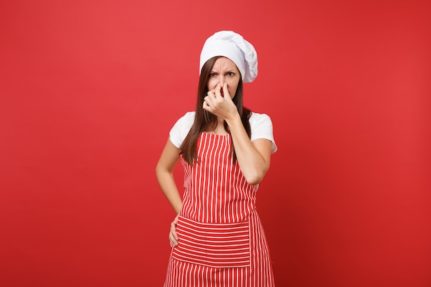 Dona de casa feminina chef cozinheira ou padeiro no avental listrado, camiseta branca, chapéu de chefs toque isolado no fundo da parede vermelha. Mulher governanta fechar o nariz com a mão por causa do cheiro. Mock-se o conceito de espaço de cópia.