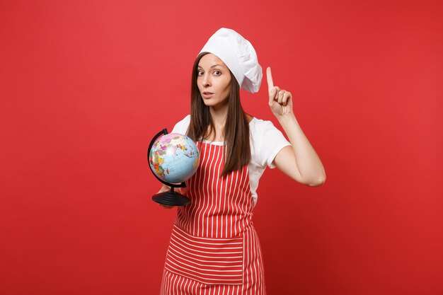 Dona de casa feminina chef cozinheira ou padeiro no avental listrado, camiseta branca, chapéu de chefs toque isolado no fundo da parede vermelha. Mulher governanta segurando nas palmas do globo do mundo da terra. Mock-se o conceito de espaço de cópia.