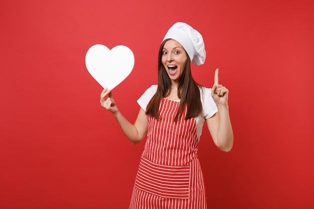 Foto dona de casa feminina chef cozinheira ou padeiro no avental listrado, camiseta branca, chapéu de chefs toque isolado no fundo da parede vermelha. mulher sorridente dona de casa segurar o espaço de trabalho de coração em branco. mock-se o conceito de espaço de cópia.