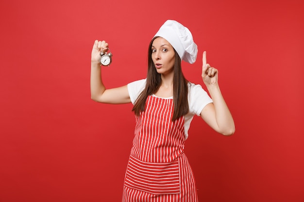 Dona de casa feminina chef cozinheira ou padeiro no avental listrado, camiseta branca, chapéu de chefs toque isolado no fundo da parede vermelha. Uma linda mulher segura na mão retrô despertador se apresse. Mock-se o conceito de espaço de cópia.