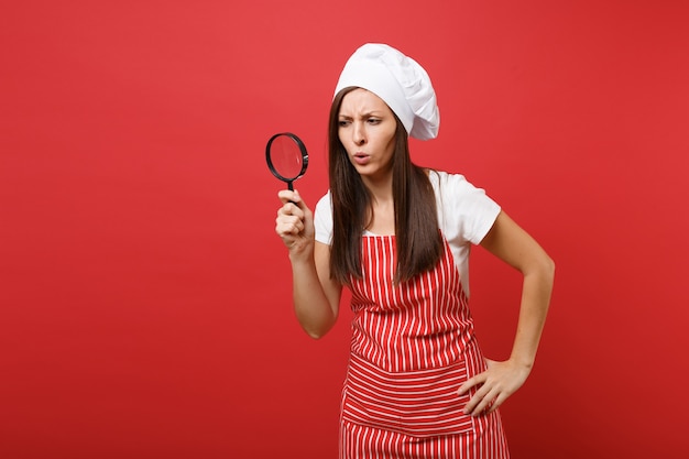 Dona de casa feminina chef cozinheira ou padeiro no chapéu de chefs avental listrado branco t-shirt toque isolado no fundo da parede vermelha. Mulher governanta segurar e olhar através da lupa. Simule o conceito de espaço de cópia