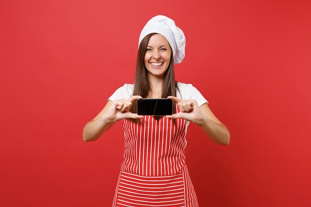 Dona de casa feminina chef cozinheira ou padeiro no chapéu de chefs avental listrado branco t-shirt toque isolado no fundo da parede vermelha. mulher segura a tela em branco do celular para conteúdo promocional simulação do conceito de espaço de cópia