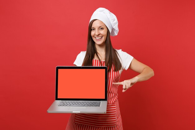 Dona de casa feminina chef cozinheira ou padeiro no chapéu de chefs avental listrado branco t-shirt toque isolado no fundo da parede vermelha. mulher segura a tela em branco do laptop para conteúdo promocional simulação do conceito de espaço de cópia