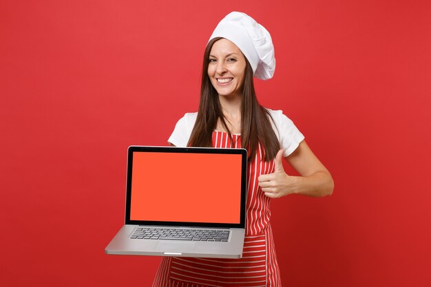 Dona de casa feminina chef cozinheira ou padeiro no chapéu de chefs avental listrado branco t-shirt toque isolado no fundo da parede vermelha. Mulher segura a tela em branco do laptop para conteúdo promocional Simulação do conceito de espaço de cópia