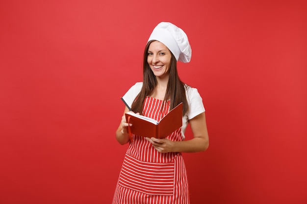 Dona de casa feminina chef cozinheira ou padeiro no chapéu de chefs avental listrado branco t-shirt toque isolado no fundo da parede vermelha. Mulher sorridente segura caneta e livro de culinária de receita do bloco de notas. Mock-se o conceito de espaço de cópia.