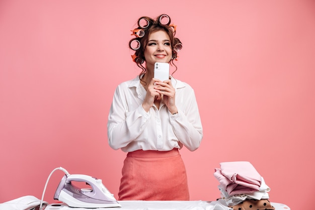 Dona de casa fofa e intrigada com rolos de cabelo segurando um telefone nas mãos em uma parede rosa