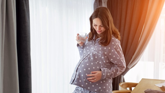 Dona de casa grávida de pijama bebe água e acaricia a barriga