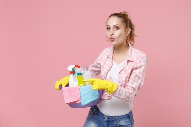 Dona de casa jovem espantada em luvas de borracha segura bacia com garrafas de detergente lavando produtos de limpeza fazendo trabalhos domésticos isolados no estúdio de fundo rosa. Conceito de limpeza. Apontando o dedo indicador para o lado.