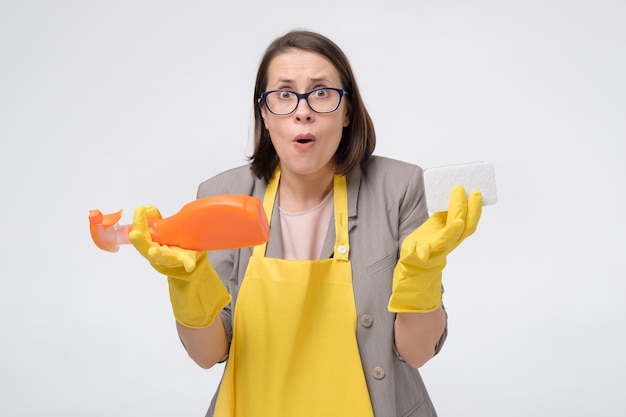 Foto dona de casa jovem estressada em um avental amarelo com esponja de cozinha e uma garrafa de detergente