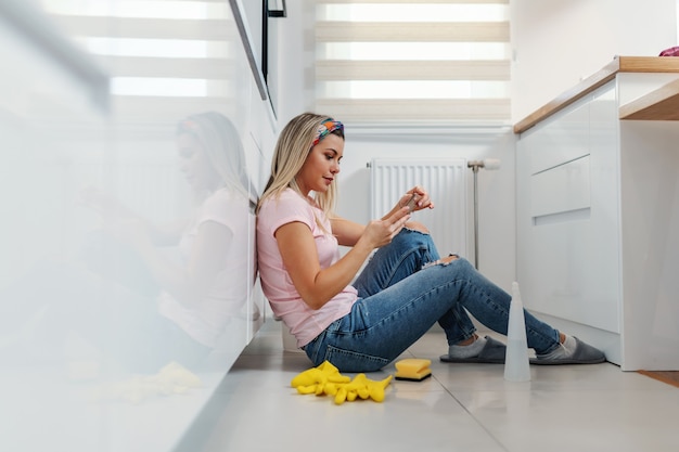Dona de casa loira digna cansada sentada no chão da cozinha, fazendo uma pausa e se divertindo em sites de mídia social.