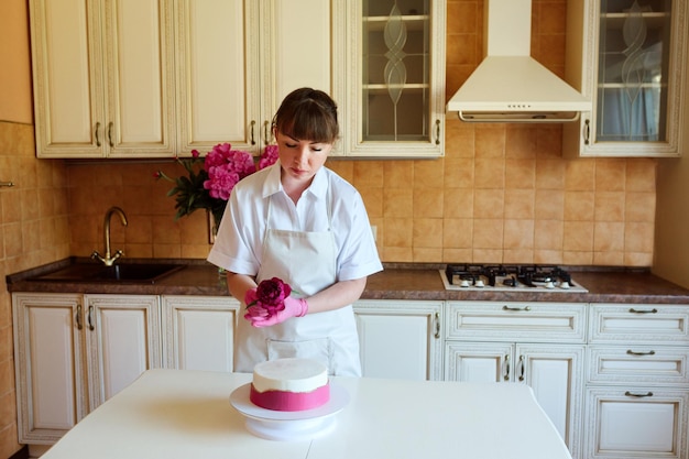 Dona de casa ou chef confeiteiro decorando delicioso bolo de aniversário ou casamento com flores de peônia em casa dentro de casa na cozinha