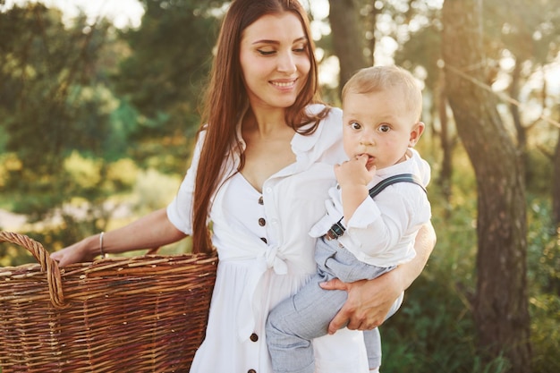 Dona de casa positiva com roupas brancas para secar Jovem mãe com seu filho pequeno está ao ar livre na floresta Lindo sol