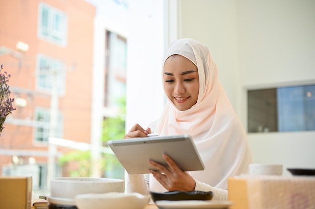 Dona de loja online muçulmana asiática muito jovem respondendo a pergunta do cliente via tablet