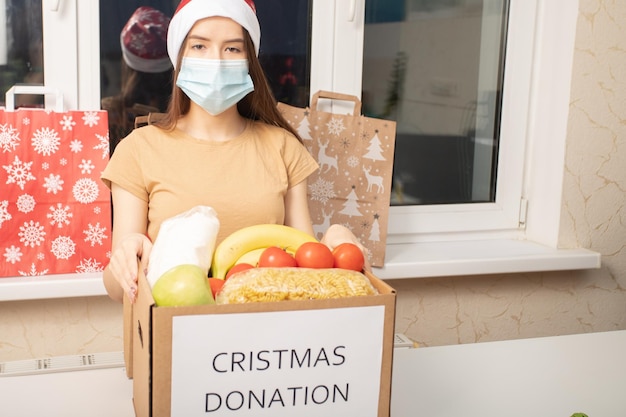 Foto donación de covid de navidad una joven voluntaria que usa una máscara médica en un centro de caridad recolecta alimentos en una caja para las víctimas de la pandemia del coronavirus