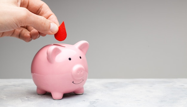 Donador de sangre. Un hombre sostiene una gota roja de sangre en su mano y la pone en una alcancía como donación. Fondo gris