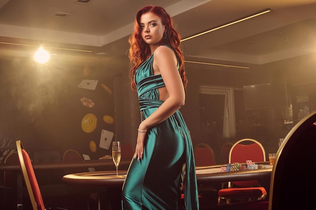 Una doncella pelirroja sexy con un vestido largo de satén azul posa de lado contra una mesa de póquer y mira la cámara en un casino de lujo. Pasión, cartas, fichas, alcohol, ganar, apostar: es como una mujer en