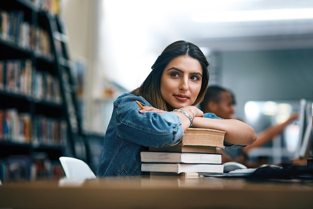 Donde los sueños se hacen realidad Foto de una mujer joven descansando sobre una pila de libros en la biblioteca de una universidad y luciendo pensativa