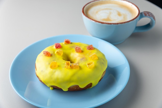 Donut coberto com esmalte amarelo e marmelada multicolorida na placa azul e xícara de cappuccino