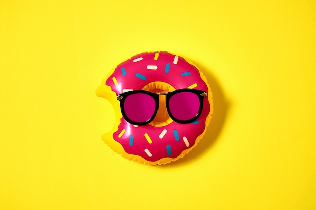 Donut inflable en gafas de sol rosas sobre fondo amarillo. Concepto mínimo creativo. Vista superior endecha plana