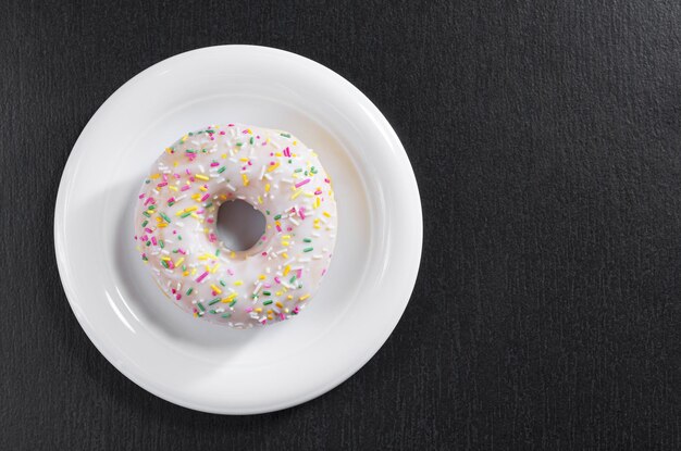 Donut mit bunten Streuseln und Zuckerguss in der Platte auf schwarzem Steinhintergrund, Draufsicht