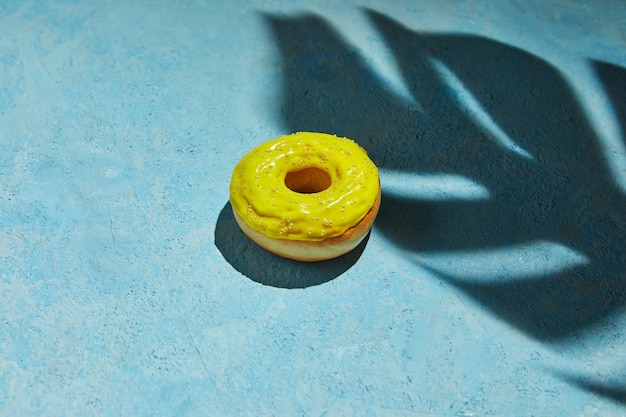 Donut mit gelbem Zuckerguss und Streuseln mit Schatten vom Monsterblatt auf blauem Hintergrund.