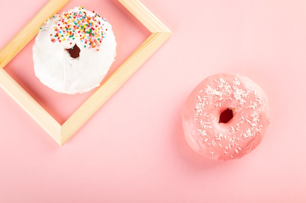 Donut mit weißem und rosa Zuckerguss und mehrfarbigem Zuckergebäck auf rosa Hintergrund. Ein Donut im Holzrahmen.