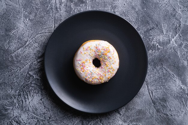 Donut de vainilla con chispas en placa negra, postre dulce glaseado sobre mesa con textura de hormigón, vista superior