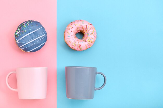 Donuts con glaseado y tazas de café sobre superficie rosa y azul pastel